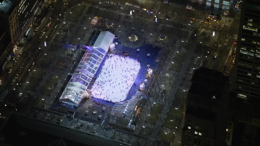 Aerial footage orbits a skyscraper in Midtown Manhattan at night to reveal a bustling ice skating rink at Bryant Park