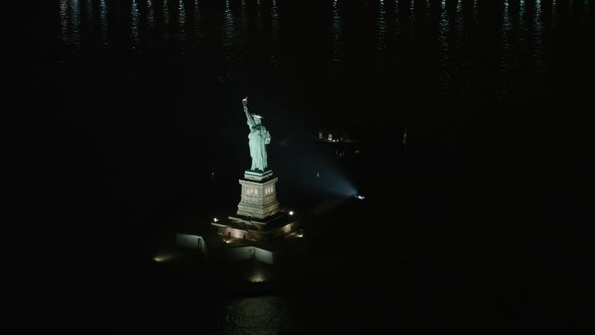 A slow approach to the iconic Statue of Liberty in New York showcases the historic landmark from a birds eye view at night with spotlights shining on the monument