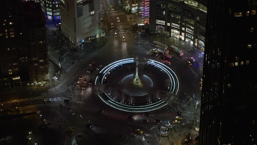 Police vehicles with flashing lights are situated around a round ornamental structure as light traffic flows during winter night in Columbus Circle Midtown Manhattan
