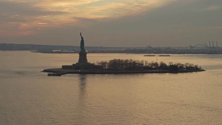 Aerial footage of Statue of Liberty during winter sunset showcasing its iconic structure on Liberty Island amidst cloudy skies and the historic harbor of New York City