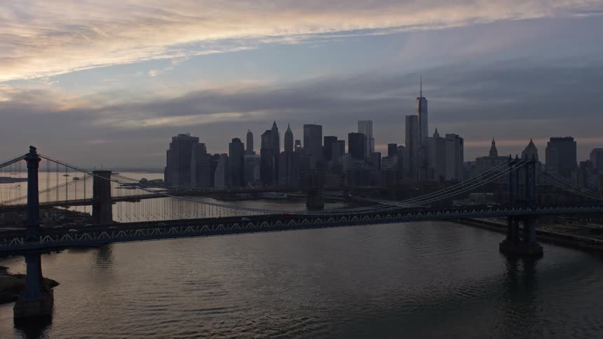 Aerial view flying over the Manhattan Bridge and approaching the historic landmark of the Brooklyn Bridge and Lower Manhattan skyline during twilight on a cloudy winter day in New York City with traffic crossing the bridges