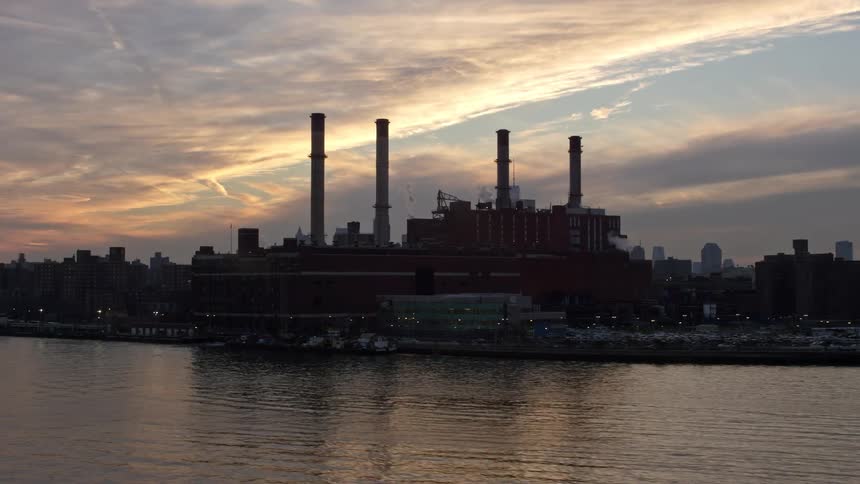 A low flying aerial view of a power plant on the waterfront of Lower Manhattan with smokestacks and the New York skyline in the distance at sunset during winter without snow