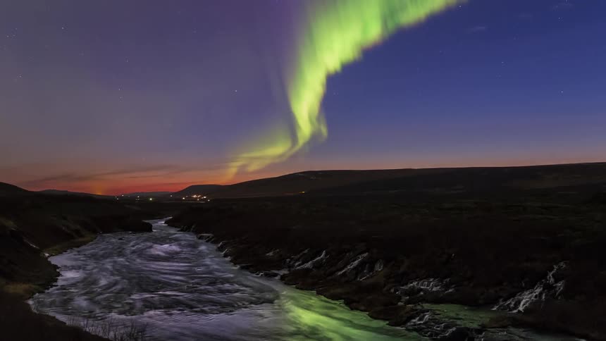 Green polar lights floating and shimmering over a river near a town at evening