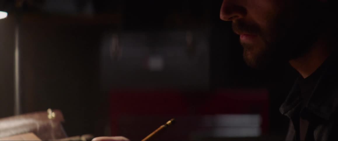 A man writes while sitting at a desk in a dark room