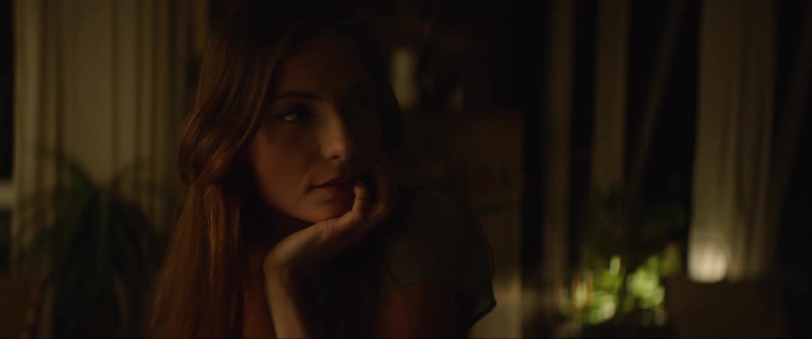 A woman is sitting in a dimly lit room resting her chin on her hand