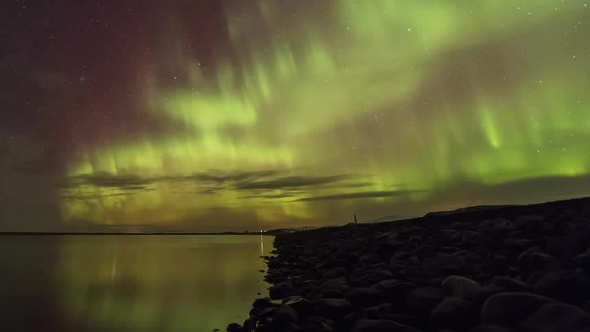 The bright green glow of the aurora borealis moves across the starry night sky along the coast