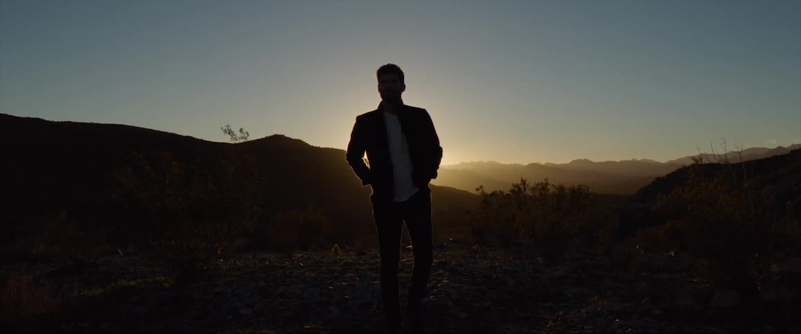 A man stands and looks over a range of mountains as the sun hangs low in the sky