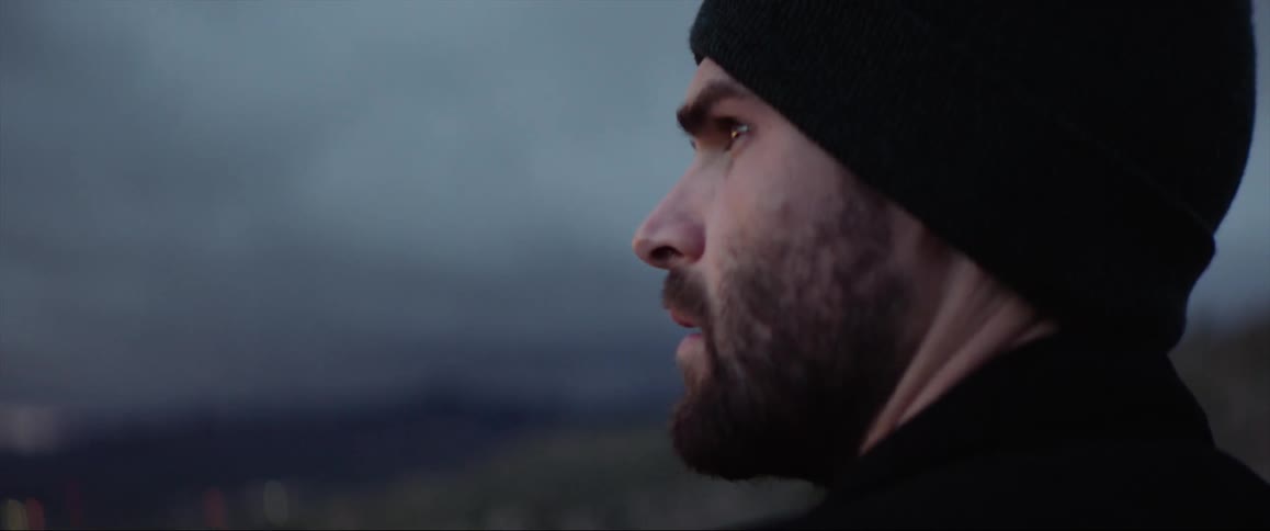 Man with beard and black wooly hat looks at distant city on mountain
