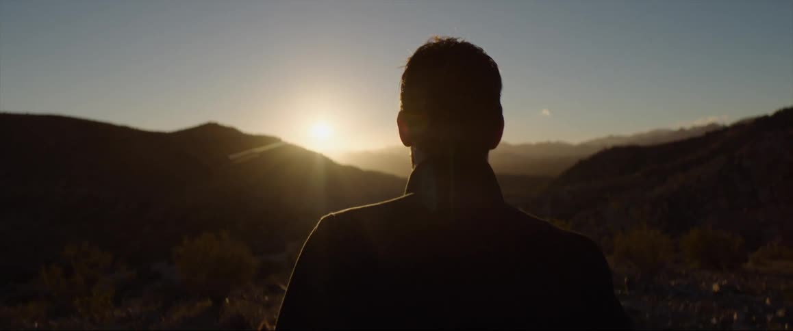 A man in a coat walks through the terrain of a hilly wilderness during the day
