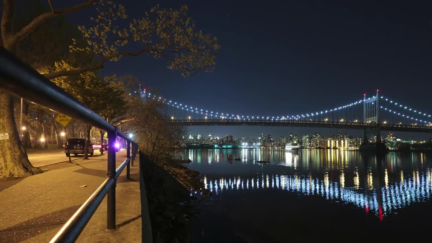 Pedestrians and traffic parallel river as ship turns under brightly lit suspension bridge at night