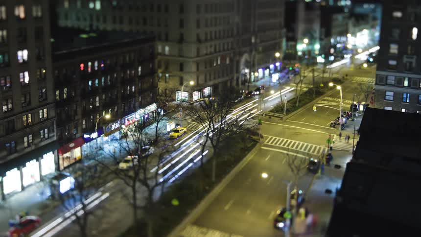 Traffic moves on a busy downtown street at night as lights are on in the buildings and people are walking on the sidewalks