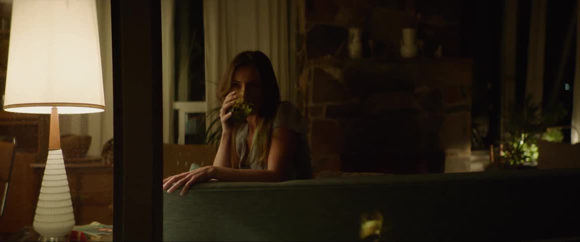 A woman holding a glass sits down on the living room couch and takes a drink