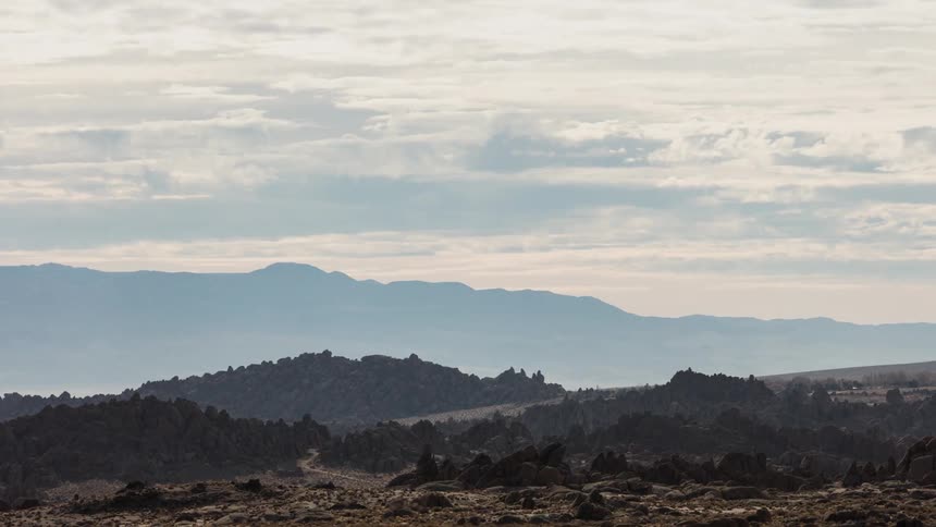 A path runs through rock formations as clouds move over mountains in a hazy sky