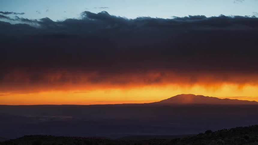 A layer of dark clouds move above a mountain over an orange sky