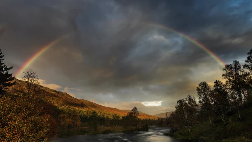 A rainbow spans a river in the countryside under a cloudy sky daytime sky