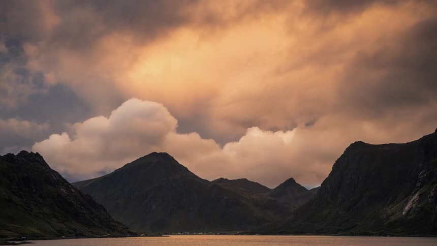 The sun shines through clouds as they move across the sky over mountains with a lake below