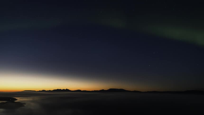 Fog moves over the water with mountains in the distance and green lights of the aurora moving across the starry sky