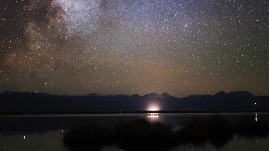 The stars of the milky way move over the mountains as traffic travels along the coast and the stars reflect on the water