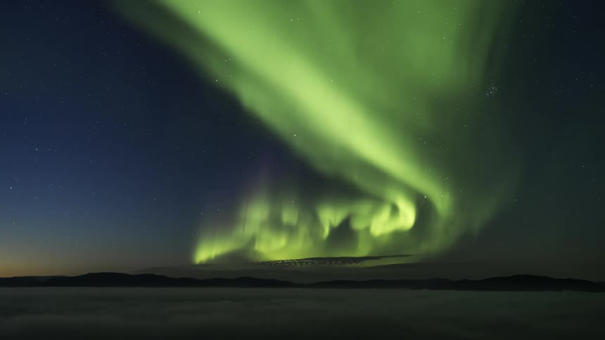 Aurora borealis dances over mountains and low lying clouds near red tinted horizon