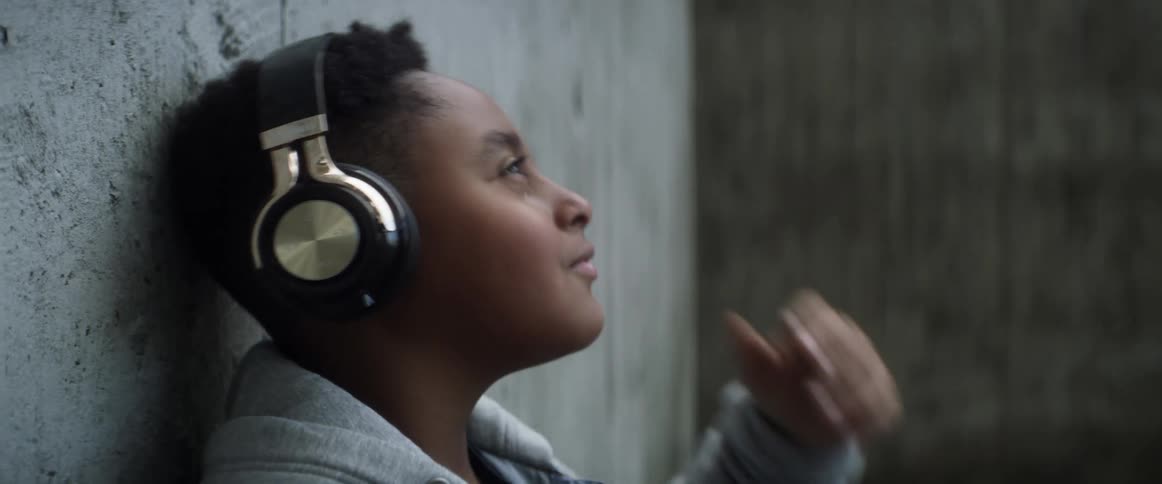 A boy is leaning against a wall with a smile on his face as he listens to headphones
