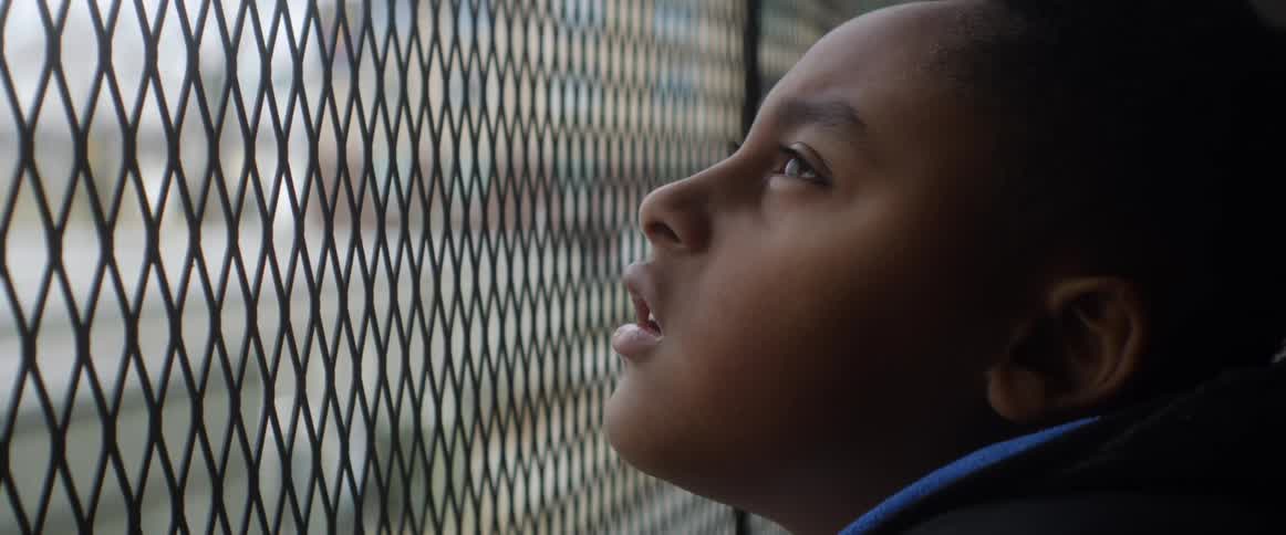 A young boy looks upwards at the sky through a metal wire fence before closing his eyes