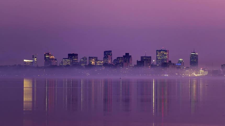 A city skyline against a darkening purple sky with lights reflecting off the water surface