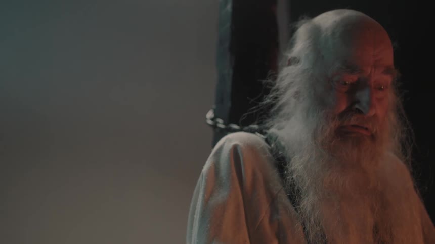 A bald man with a long white beard is chained to a wooden pole