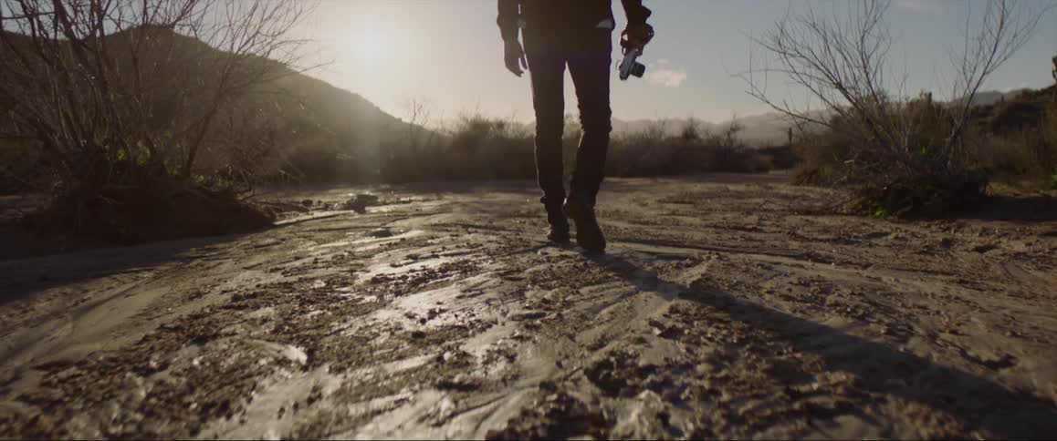 Male photographer carrying a camera moves across muddy ground between hills on a sunny day