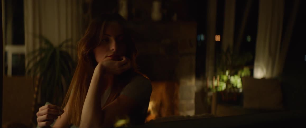 Woman gray t shirt sits on couch by fireplace and touches her hair thinking in living room at night
