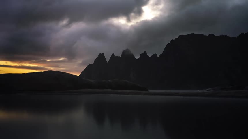 Dark clouds travel over the mountains next to the lake as the sun sets