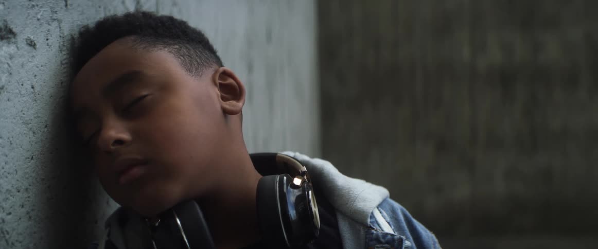 Boy with headphones and denim jacket leans against the wall and smiles slightly in basketball court