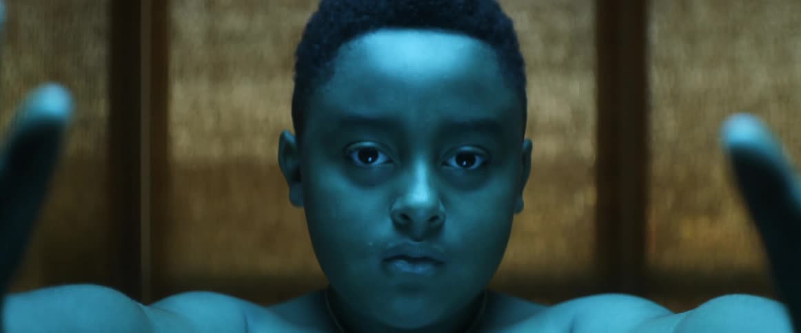Boy with blue tinted skin stands with his hands forward under colored lights