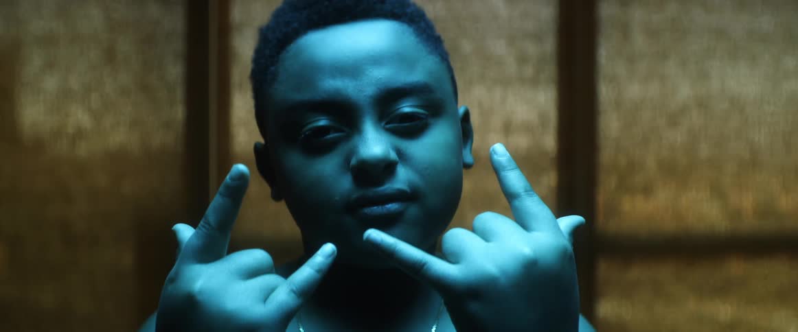 A young boy bathed in blue light poses for the camera while making hand signs