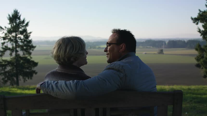A man and woman sit on a wooden bench and kiss while looking out over a field