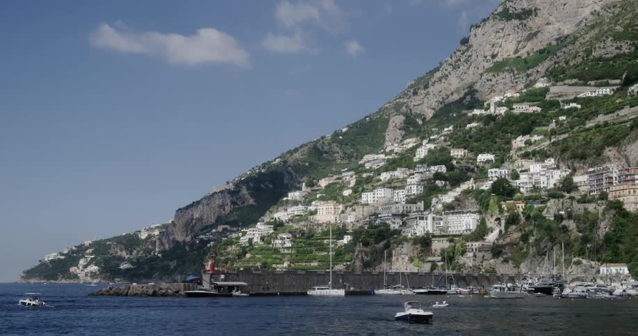 Boats move through water near docks of seaside town sitting on mountain