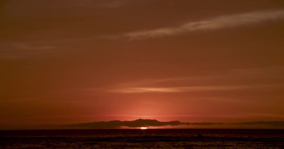 The sun sets over the water behind a cloud in a deep red sky