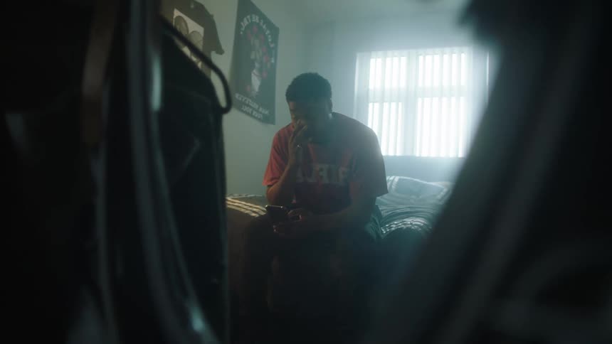 Man with orange shirt sits on bed and uses smartphone in bedroom