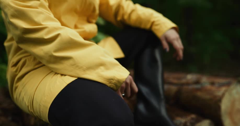 Woman wearing yellow raincoat black pants and a pair of boots sits on logs in the woods
