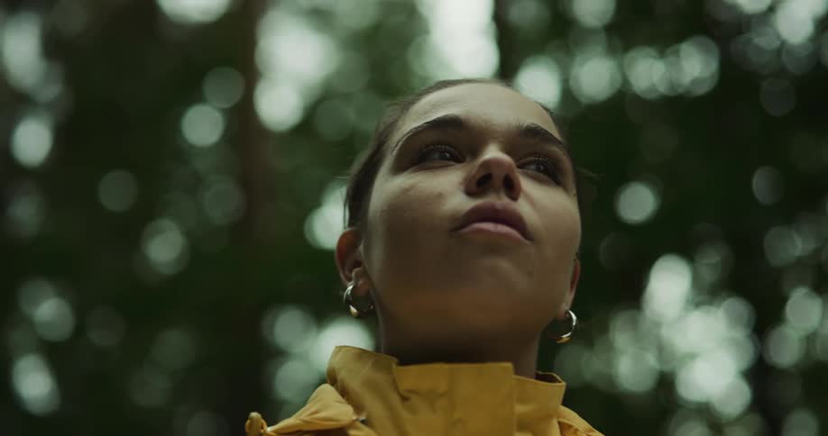 A woman in a yellow raincoat slowly looks around at the trees towering above her