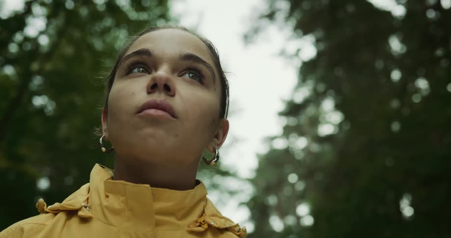 Woman with earrings and yellow raincoat looks around in the forest