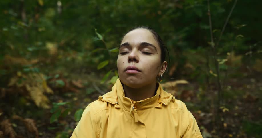 A woman in a yellow rain jacket taking deep breaths with her eyes closed in a forested area