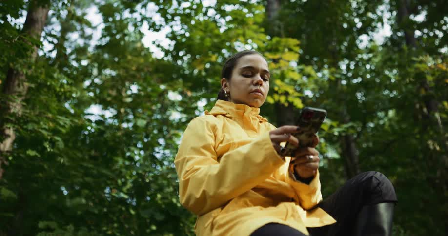 Woman wearing yellow raincoat sits outdoors among trees texting