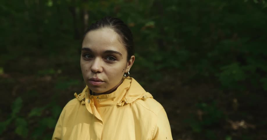 Woman with earrings and yellow raincoat stares at the camera in the woods