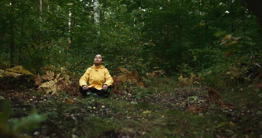 Woman in yellow raincoat sits cross legged on ground in the woods