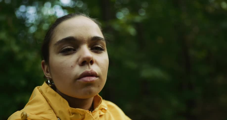 A woman in a yellow raincoat slowly turns her head to look at the camera