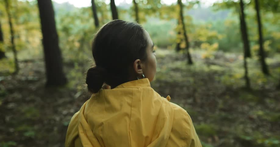 Woman with bun earrings and yellow raincoat hikes in the green woods