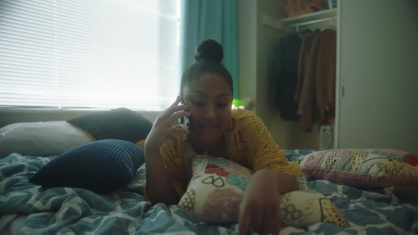 Woman with bun and yellow shirt lies in bed talking on smartphone in the bedroom