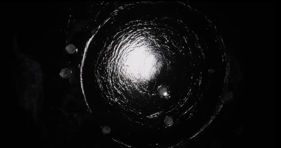 Ripples spread from bright opening through liquid strewn with pale globs