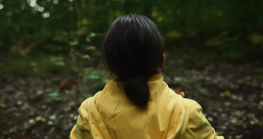 Woman with bun yellow raincoat looks up at trees in the woods