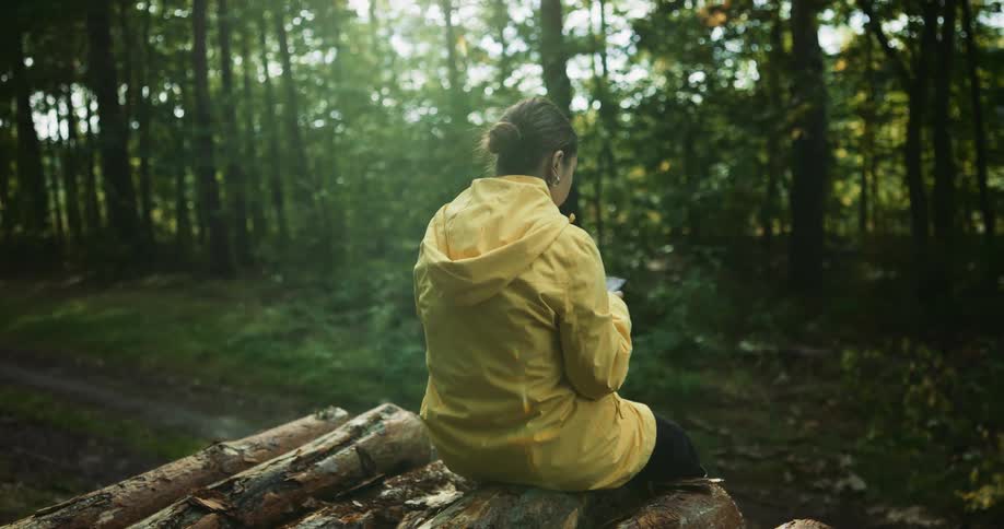 A woman sits on logs using her phone in the forest